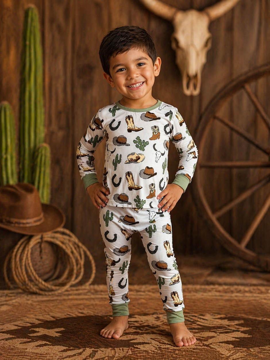Smiling child in patterned pajamas with cowboy motifs, standing on a rustic backdrop with cacti and decor.