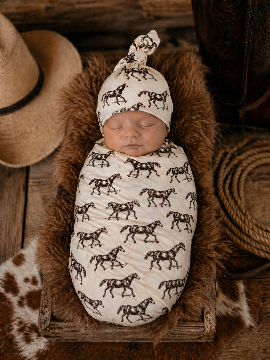 Newborn swaddled in a horse-patterned fabric, resting on a fur-textured blanket with a cowboy hat nearby.