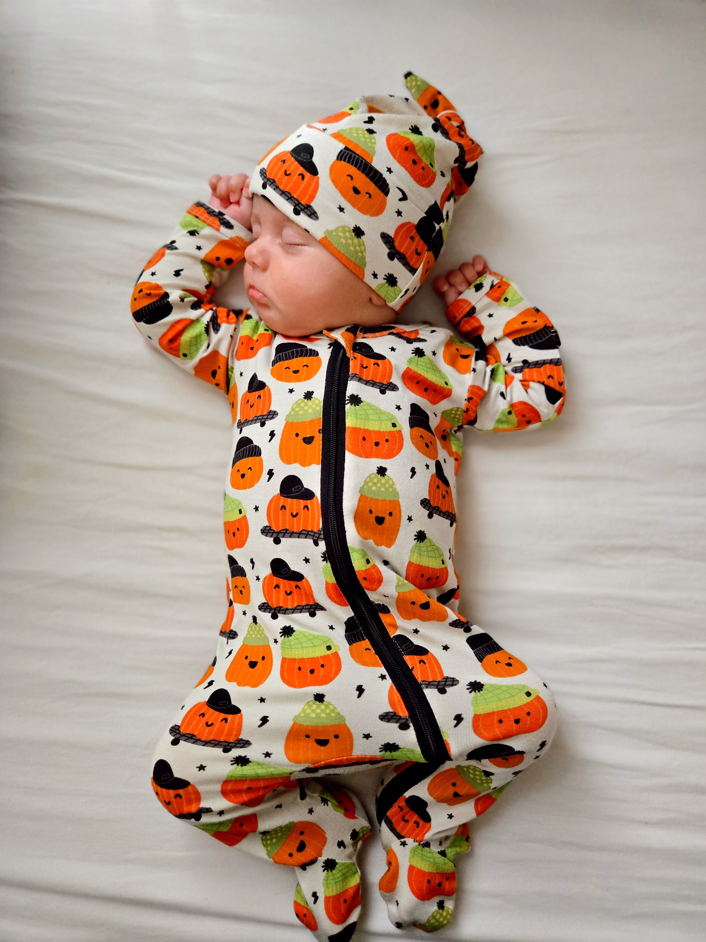 Infant sleeping on a white blanket, wearing a pumpkin-themed outfit and matching hat.
