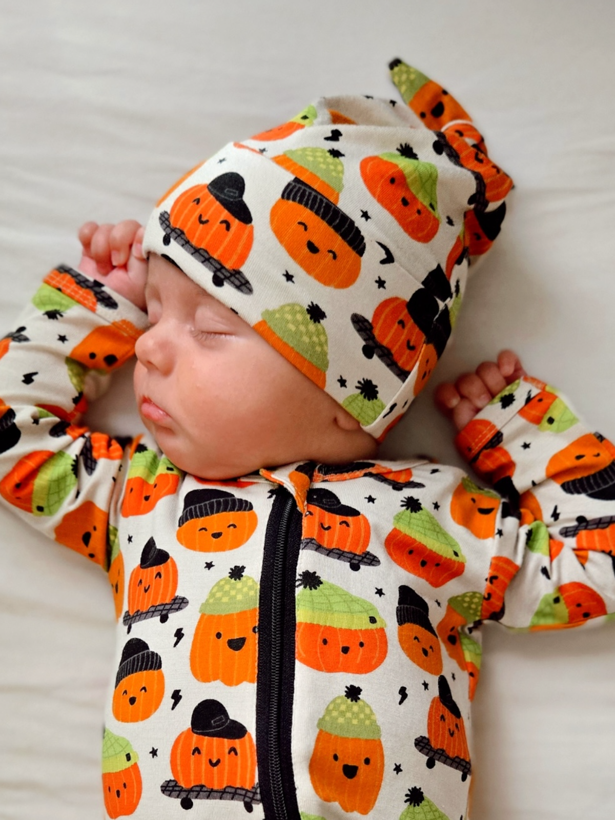 Sleeping baby in pumpkin-patterned outfit with a matching hat, resting on a light-colored surface.