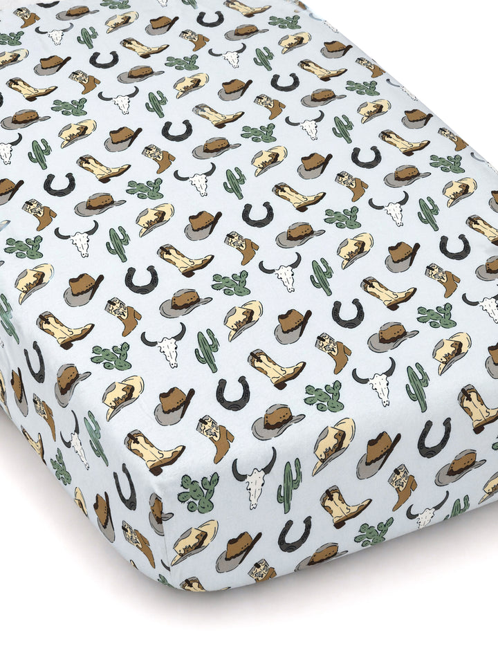 Light blue crib sheet featuring a western pattern of boots, hats, cacti, horseshoes, and cow skulls.
