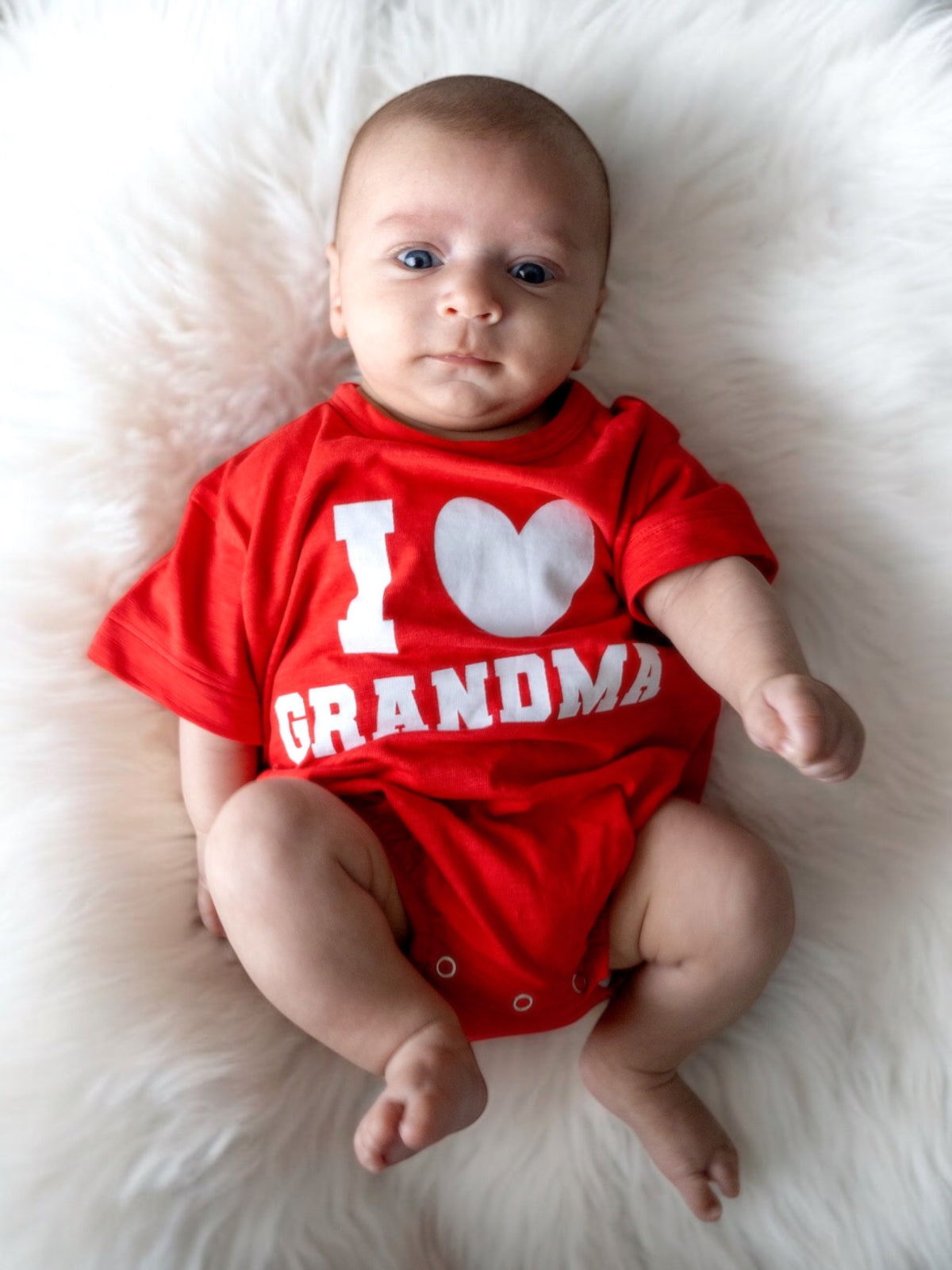 Baby wearing a red "I Love Grandma" shirt, lying on a fluffy white surface. Cute and expressive gaze.