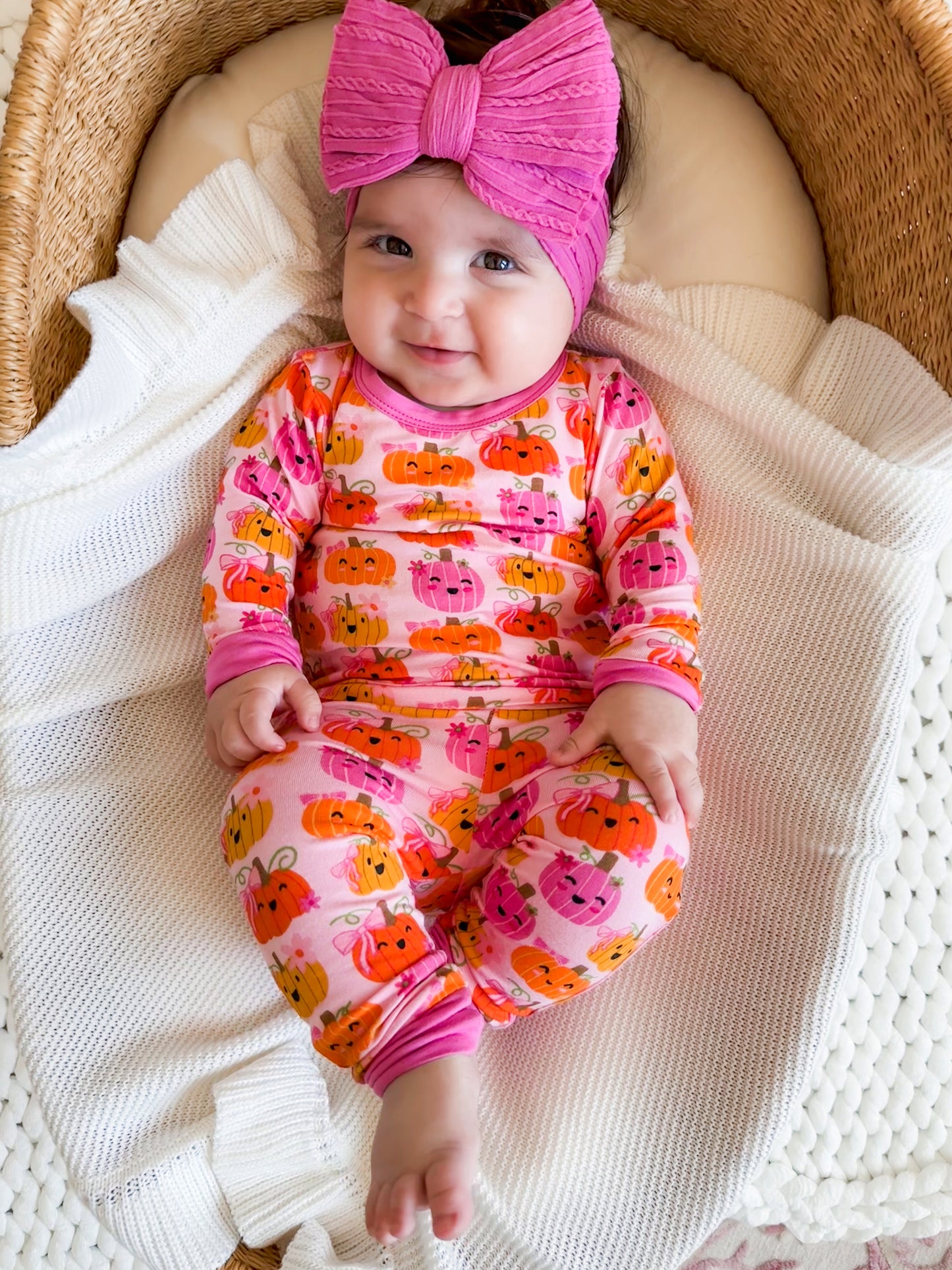 Smiling baby in a pink pumpkin-print outfit and a matching headband, sitting on a cozy blanket in a woven basket.