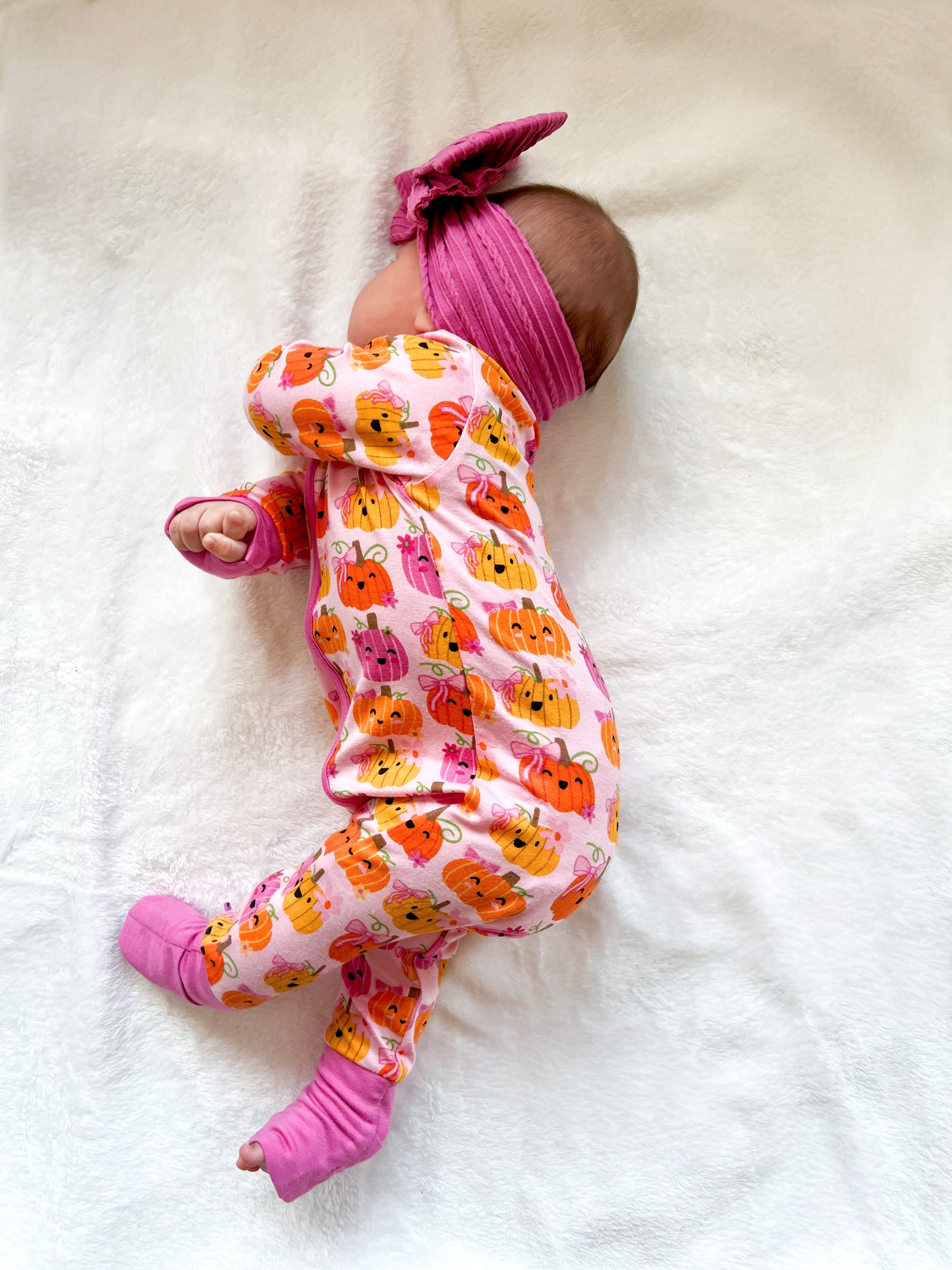 Baby girl in pumpkin-patterned pajamas and a pink headband, sleeping on a soft white blanket.