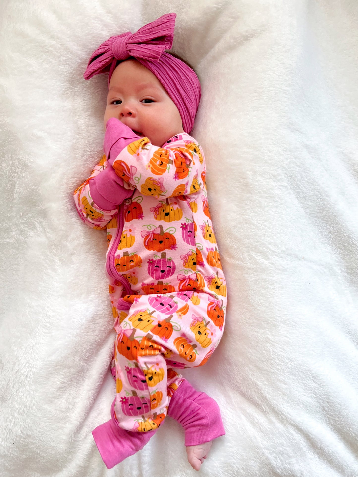 Baby in a pink pumpkin-patterned onesie with a matching pink headband, resting on a soft white blanket.