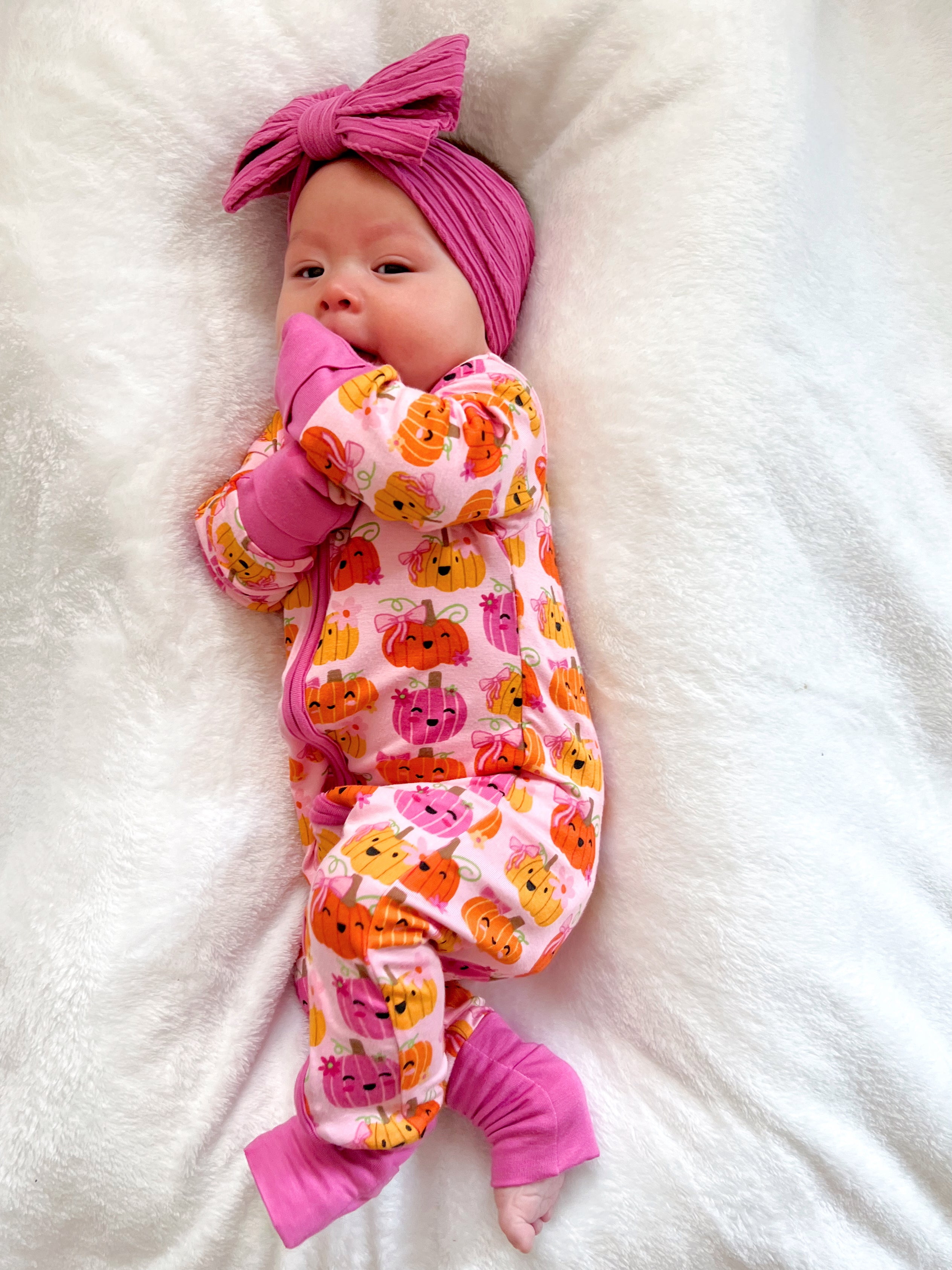 Baby in a pink pumpkin-patterned onesie with a matching pink headband, resting on a soft white blanket.