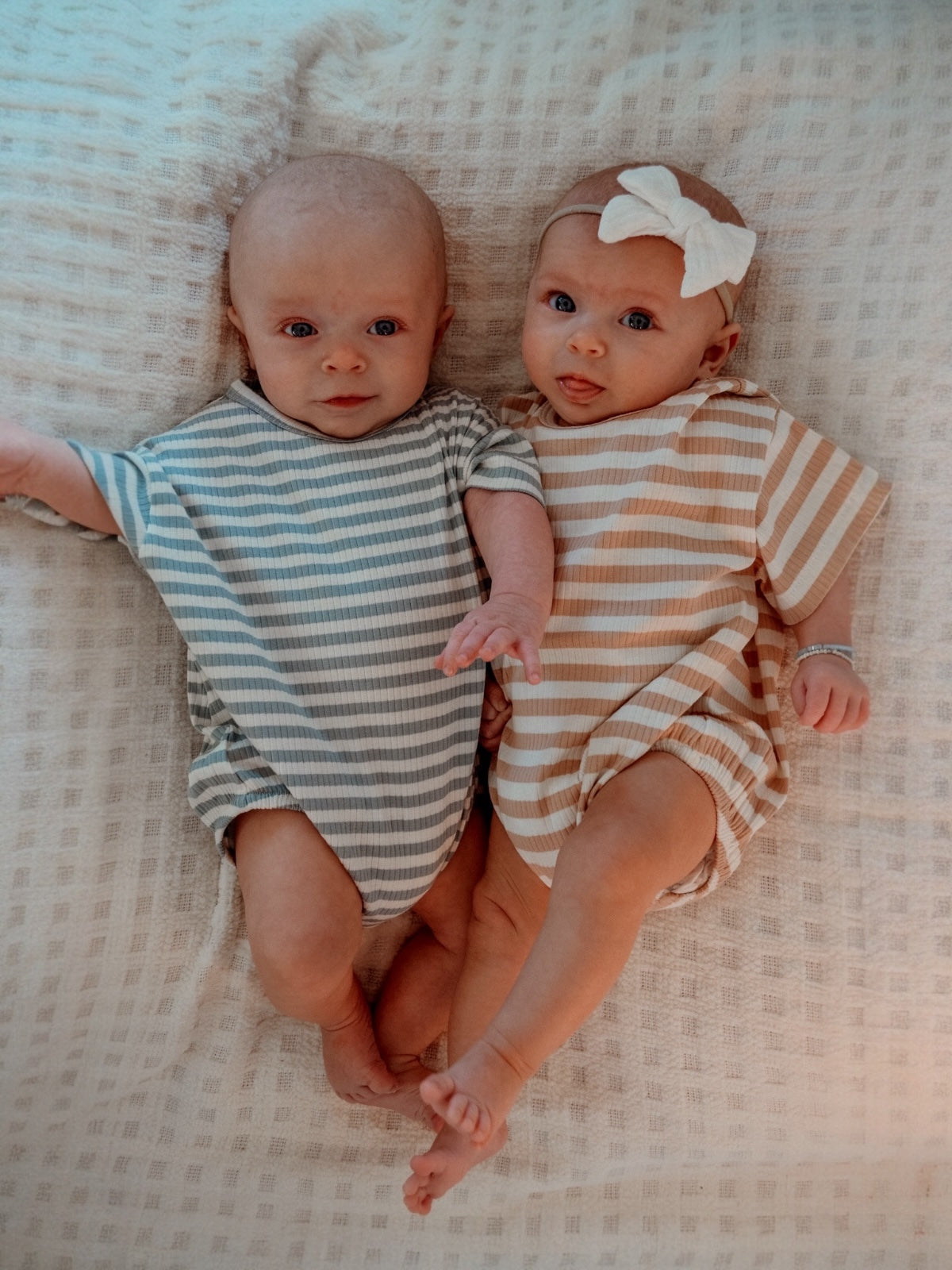 Two babies in striped romper outfits lay on a textured blanket, one with a bow in their hair.