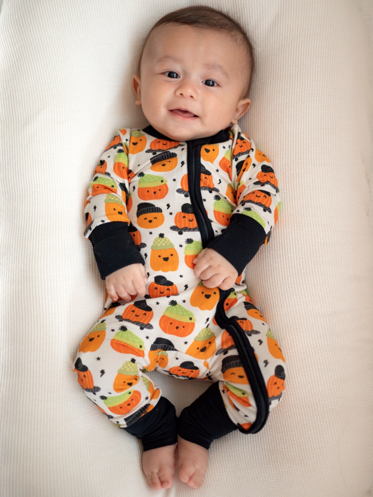 Baby in cozy outfit with colorful pumpkin print, smiling while laying on a soft, textured surface.