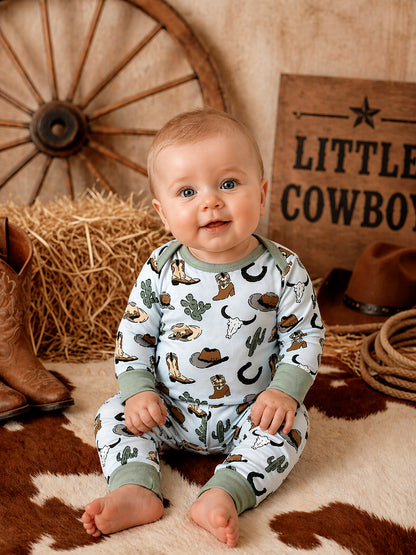 Smiling baby in cowboy-themed pajamas sits on rustic backdrop with straw, cowboy boots, and western decor.