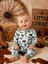 Smiling baby in cowboy-themed pajamas sits on rustic backdrop with straw, cowboy boots, and western decor.