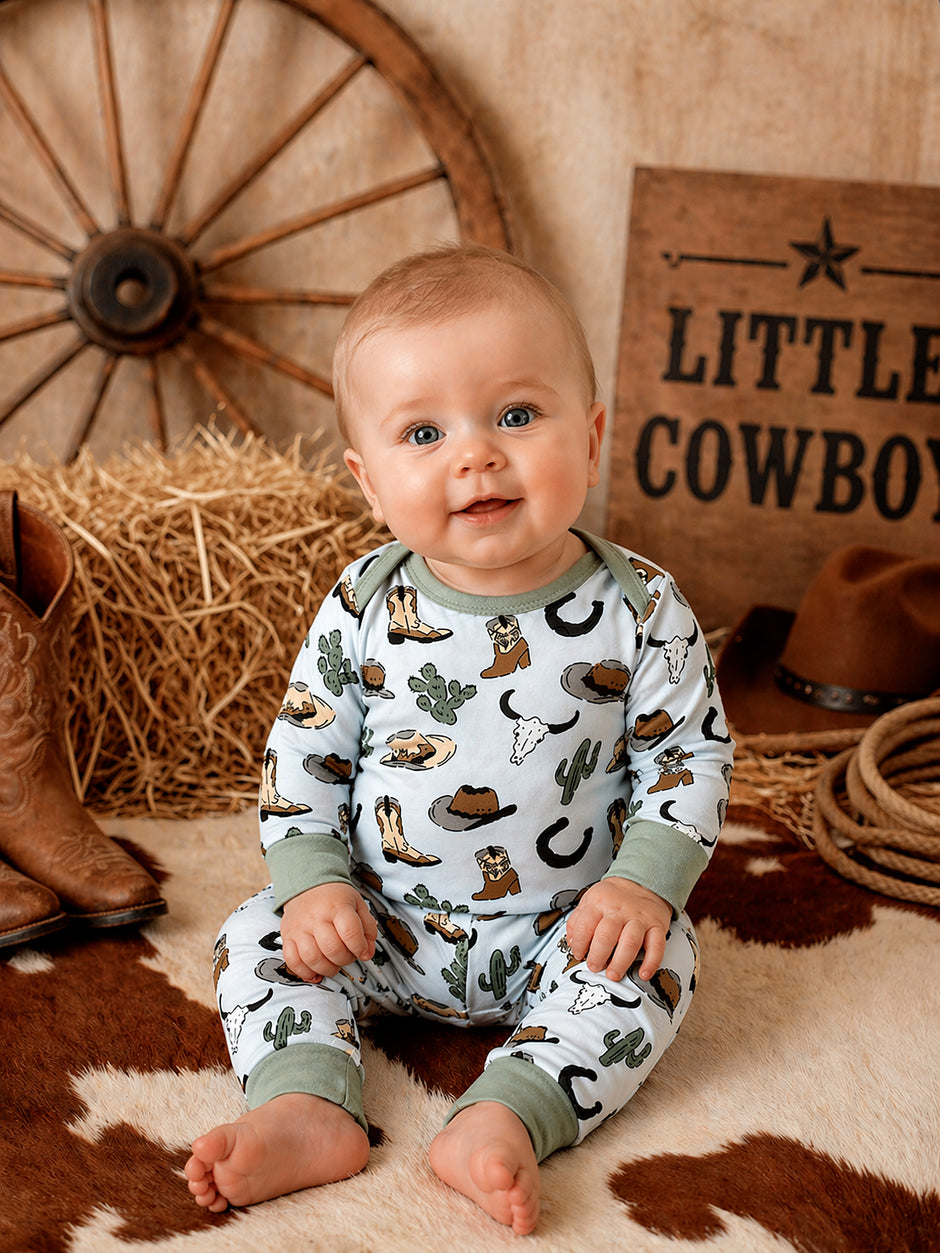 Smiling baby in cowboy-themed pajamas sits on rustic backdrop with straw, cowboy boots, and western decor.