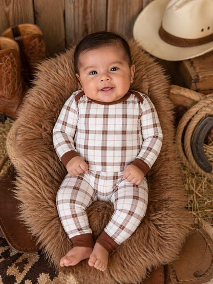 Smiling baby in plaid outfit, sitting on a fluffy rug surrounded by rustic decor, including cowboy boots and a hat.