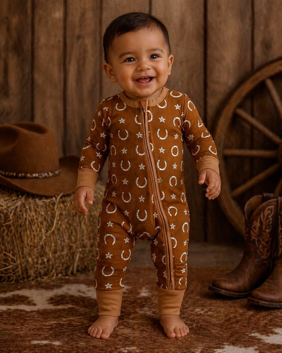 Smiling baby in a brown onesie with stars and horseshoes, standing on a cowhide rug near a cowboy hat and boots.