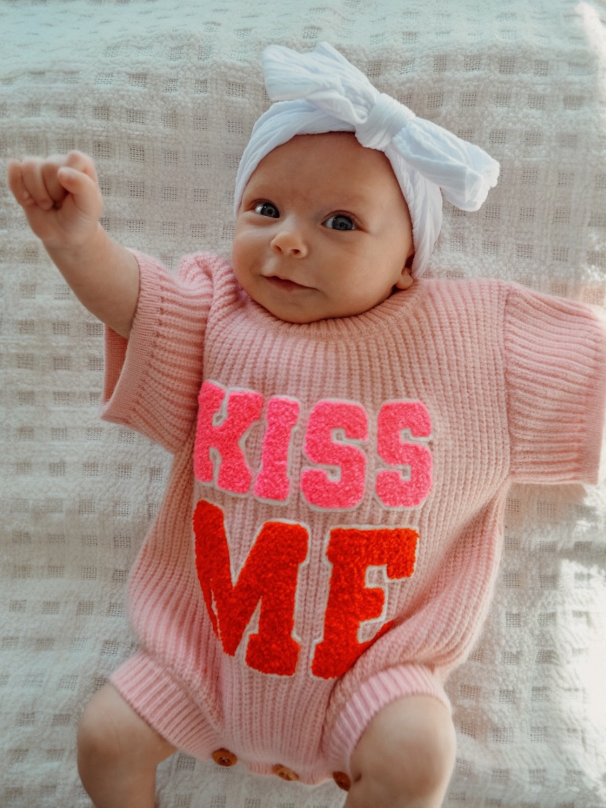 Smiling baby in a pink sweater with "KISS ME" text and a white bow headband, making a playful gesture.