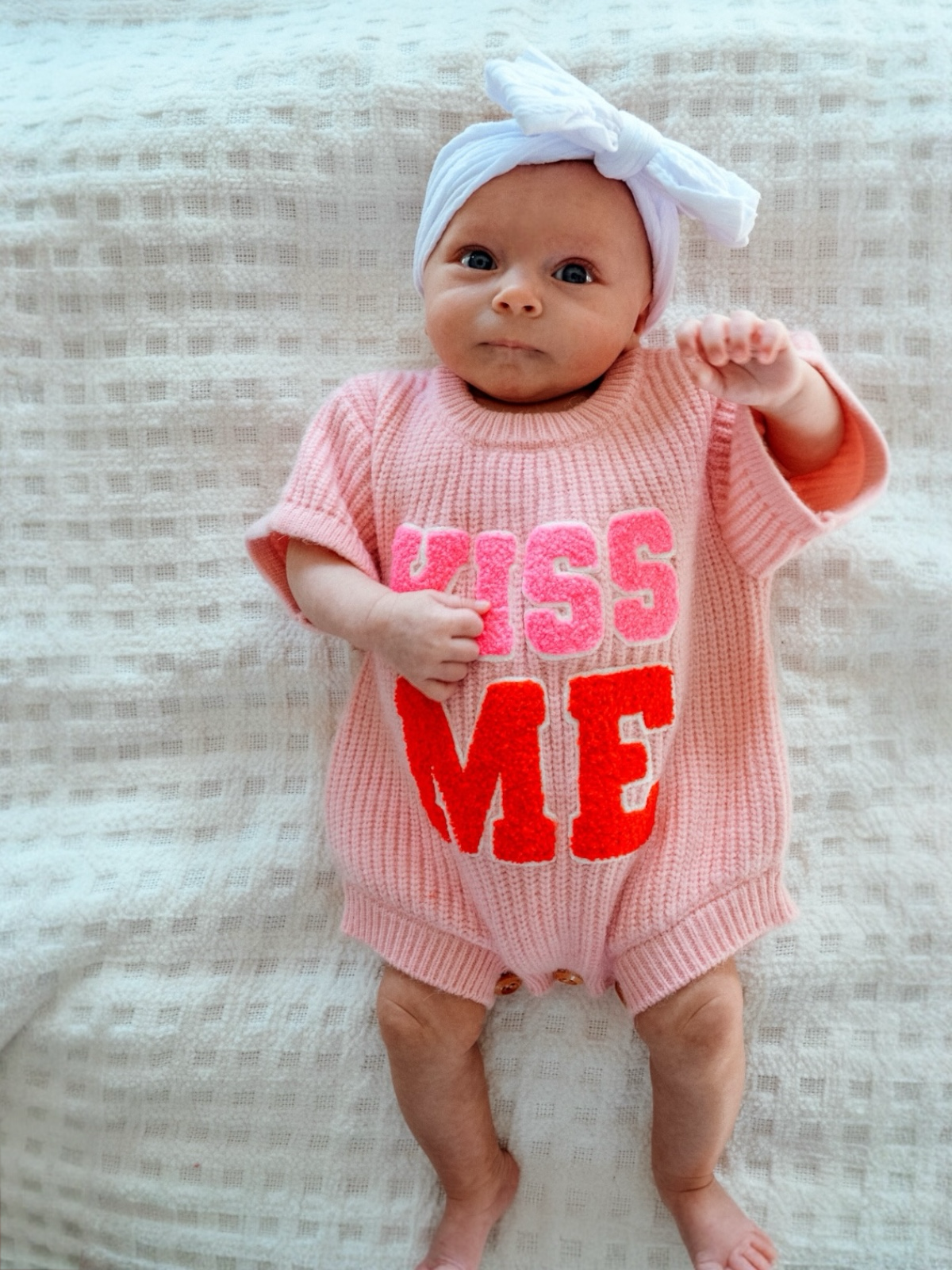 Infant in a pink outfit with "KISS ME" text, wearing a white headband, lying on a textured blanket.