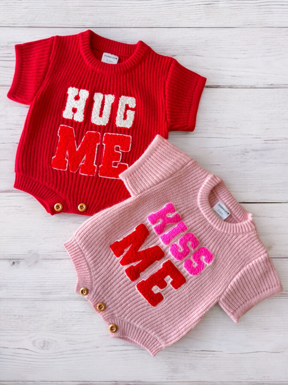 Red and pink baby rompers with "HUG ME" and "KISS ME" text on a wooden background.