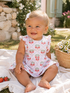 Smiling baby girl in a floral dress, sitting on a blanket with strawberries and flowers in a sunny garden setting.