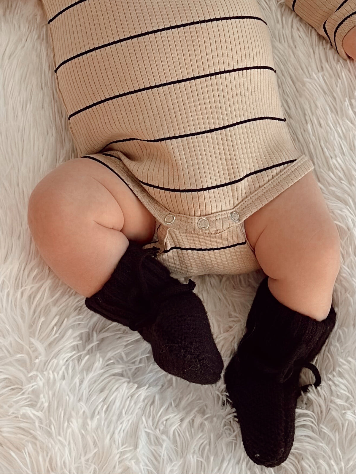 Baby's legs in a striped outfit, wearing black knitted booties, resting on a soft, fluffy white surface.