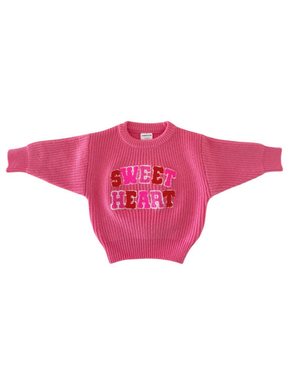 Pink sweater with "SWEET HEART" graphic in red and pink letters, featuring a ribbed texture and wide sleeves.
