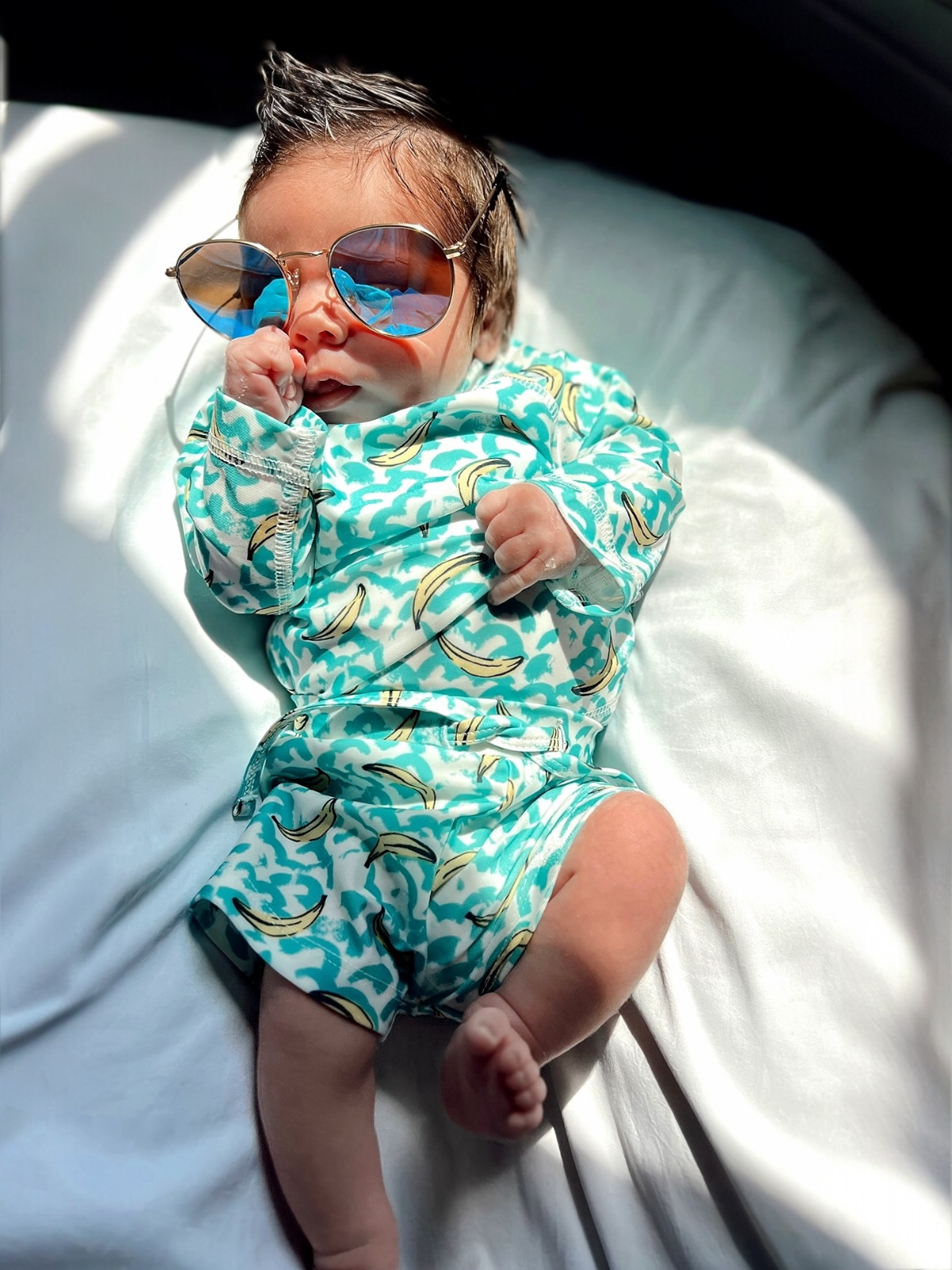 Baby dressed in a turquoise outfit with banana prints, wearing sunglasses and posing on a white background.
