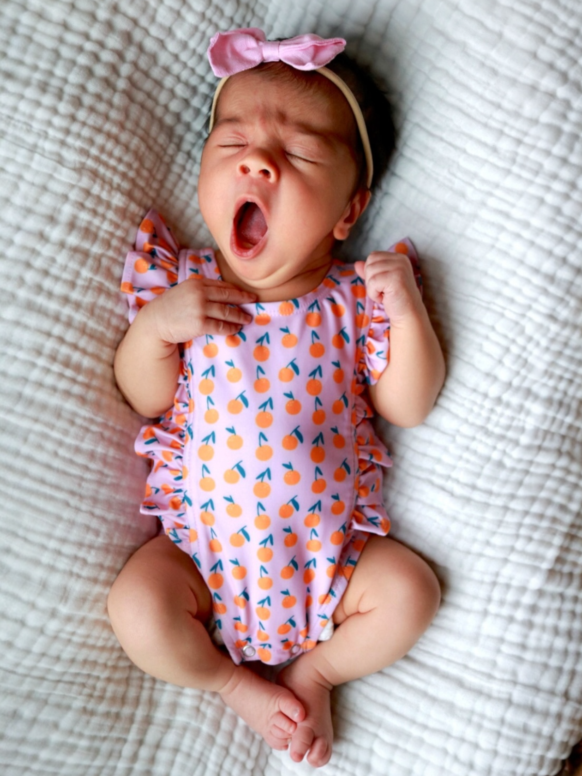 Infant yawning in a pink onesie with orange patterns, lying on a textured blanket.