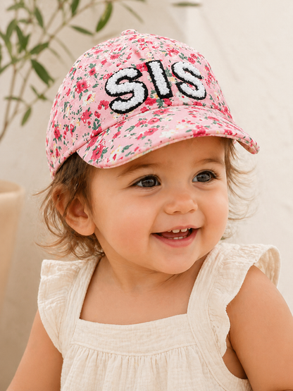 Smiling toddler wearing a pink floral cap with "SIS" embroidered on the front, set against a soft background.