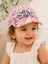 Smiling toddler wearing a pink floral cap with "SIS" embroidered on the front, set against a soft background.
