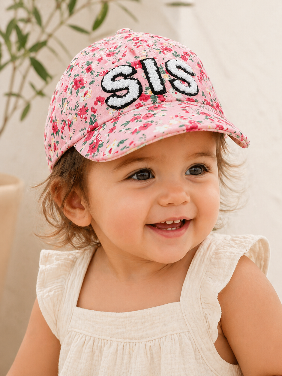 Smiling toddler wearing a pink floral cap with "SIS" embroidered on the front, set against a soft background.