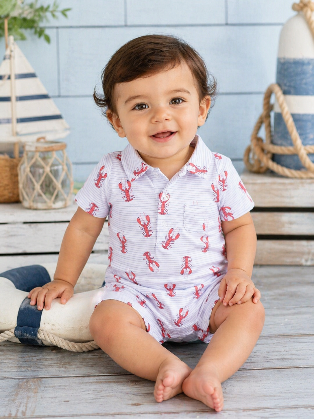 Smiling toddler in a lobster-patterned outfit, seated on a wooden floor with nautical decor in the background.