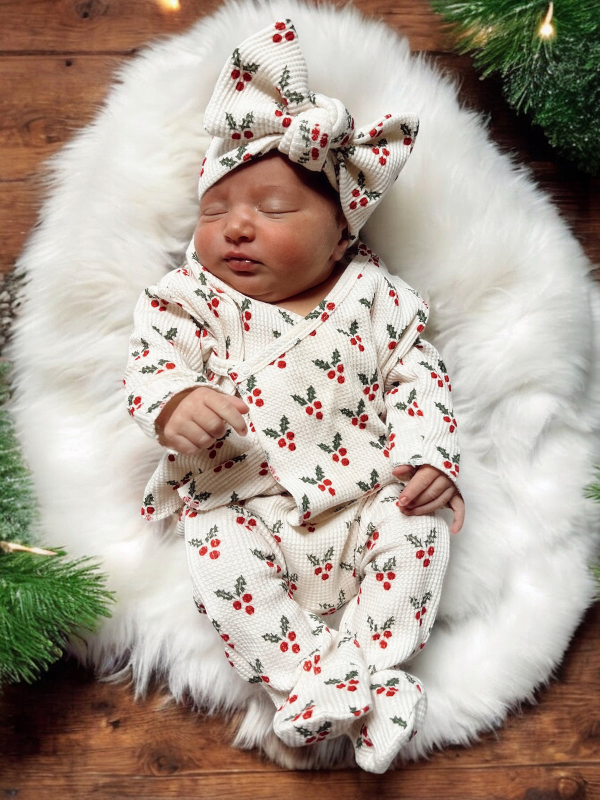 Newborn baby in holiday-themed outfit with red berries, lying on fluffy white blanket surrounded by greenery.