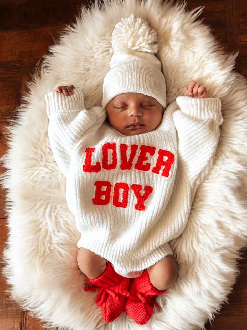 Baby wearing a white sweater with "LOVER BOY" in red, lying on a fluffy rug, with a knitted hat and red booties.
