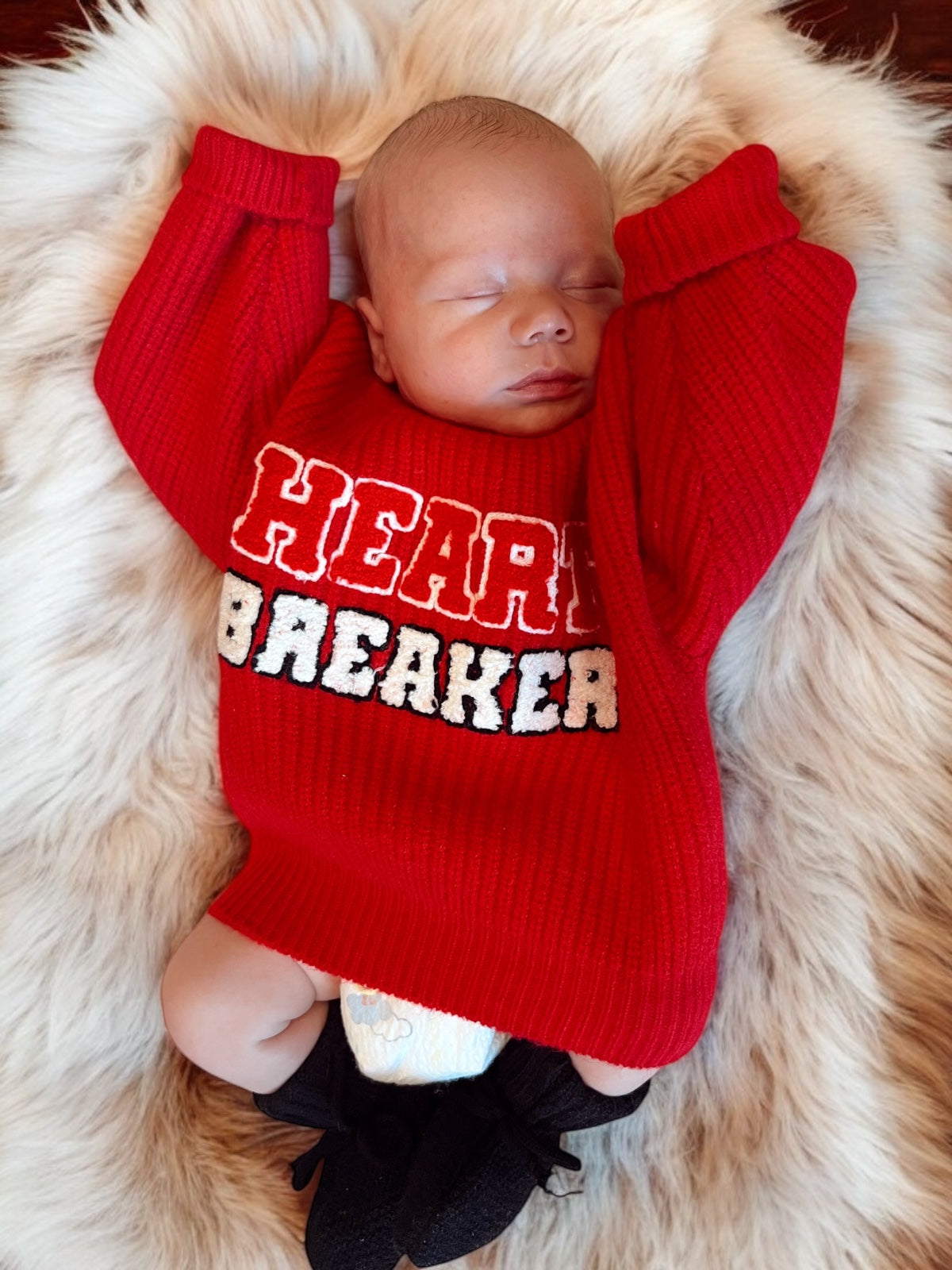 Baby in a red "Heart Breaker" sweater rests on a fluffy surface, with eyes closed and arms raised.