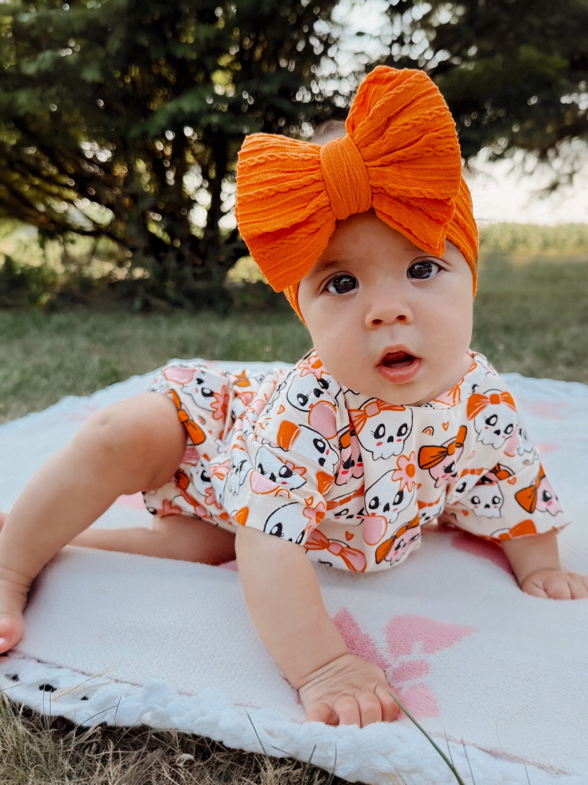 Baby sitting on a blanket outdoors, wearing a colorful bow and a playful printed outfit, surrounded by greenery.