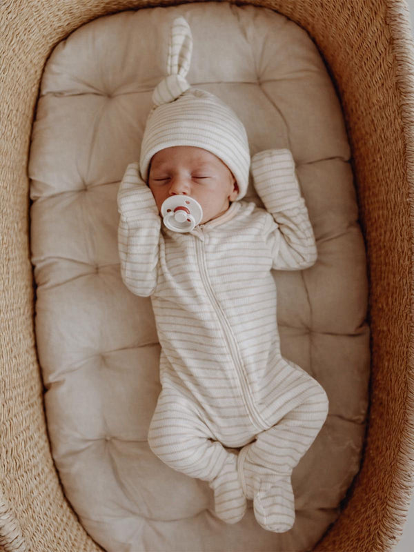 Baby in striped onesie and hat sleeps in a cozy basket, holding a pacifier on a cushioned surface.