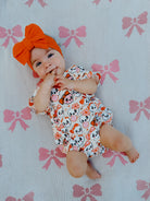 Baby with an orange bow headband, smiling on a pink bow-patterned blanket, wearing a playful animal print outfit.