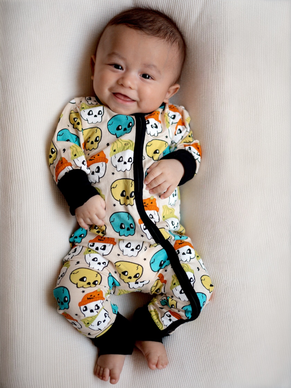Smiling baby in colorful skull-patterned pajamas, lying on a textured white surface.