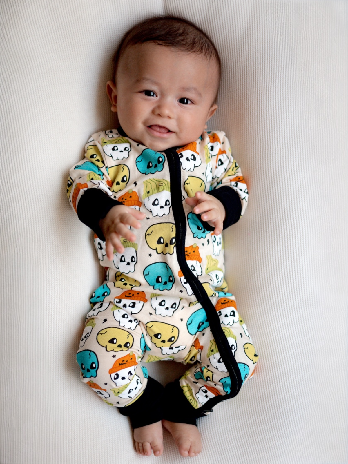 Smiling baby in colorful skull-print pajamas, sitting on a light textured surface.