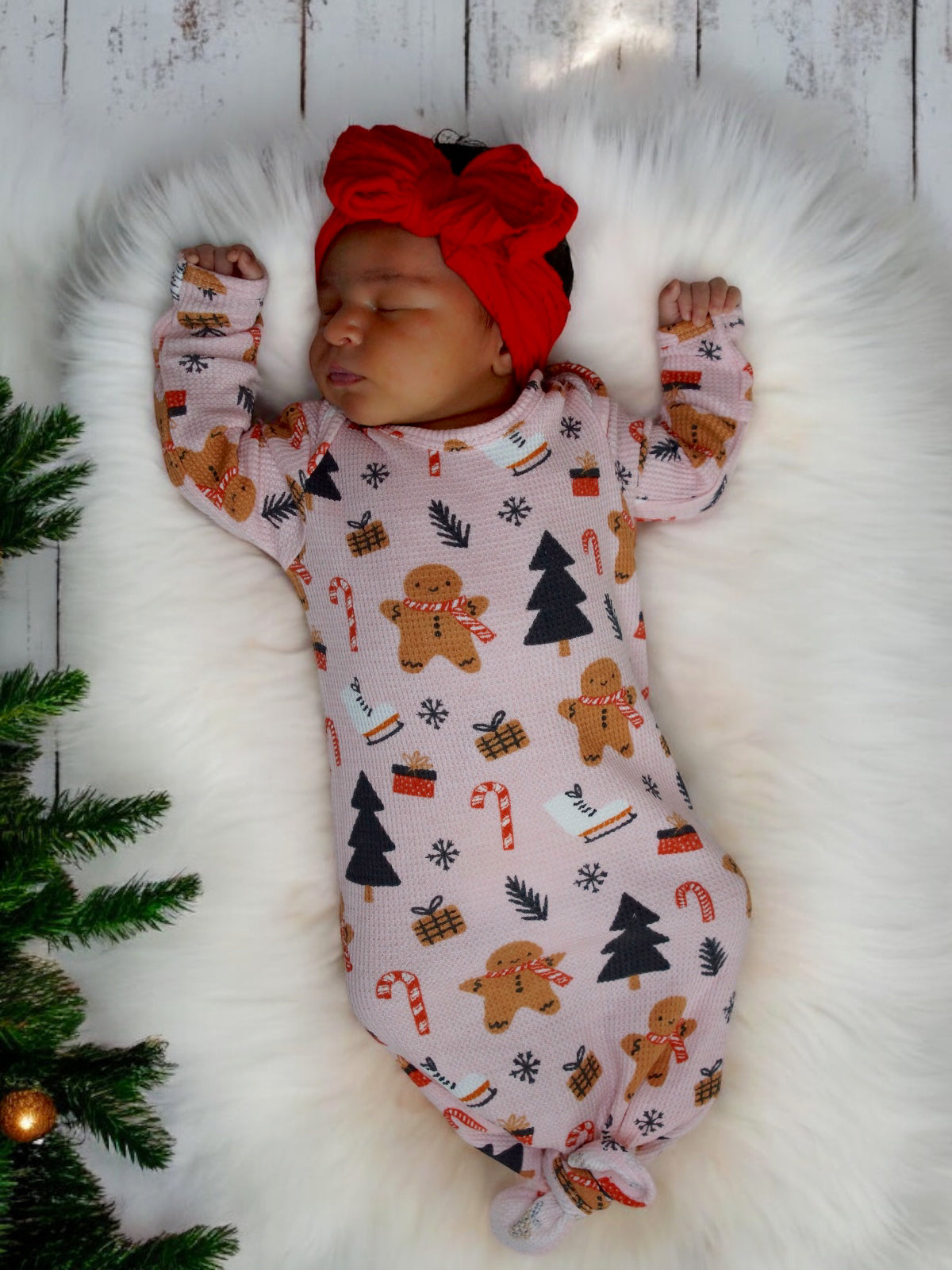 Sleeping baby in festive gingerbread onesie with red headband, surrounded by a cozy white blanket and greenery.