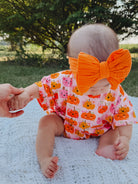 Baby sitting on a blanket outdoors, wearing a pumpkin-patterned outfit and a large orange bow, holding a hand.