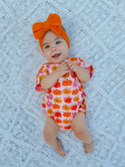 Baby lying on a white lace blanket, wearing a pumpkin-patterned outfit and an orange bow headband, smiling happily.