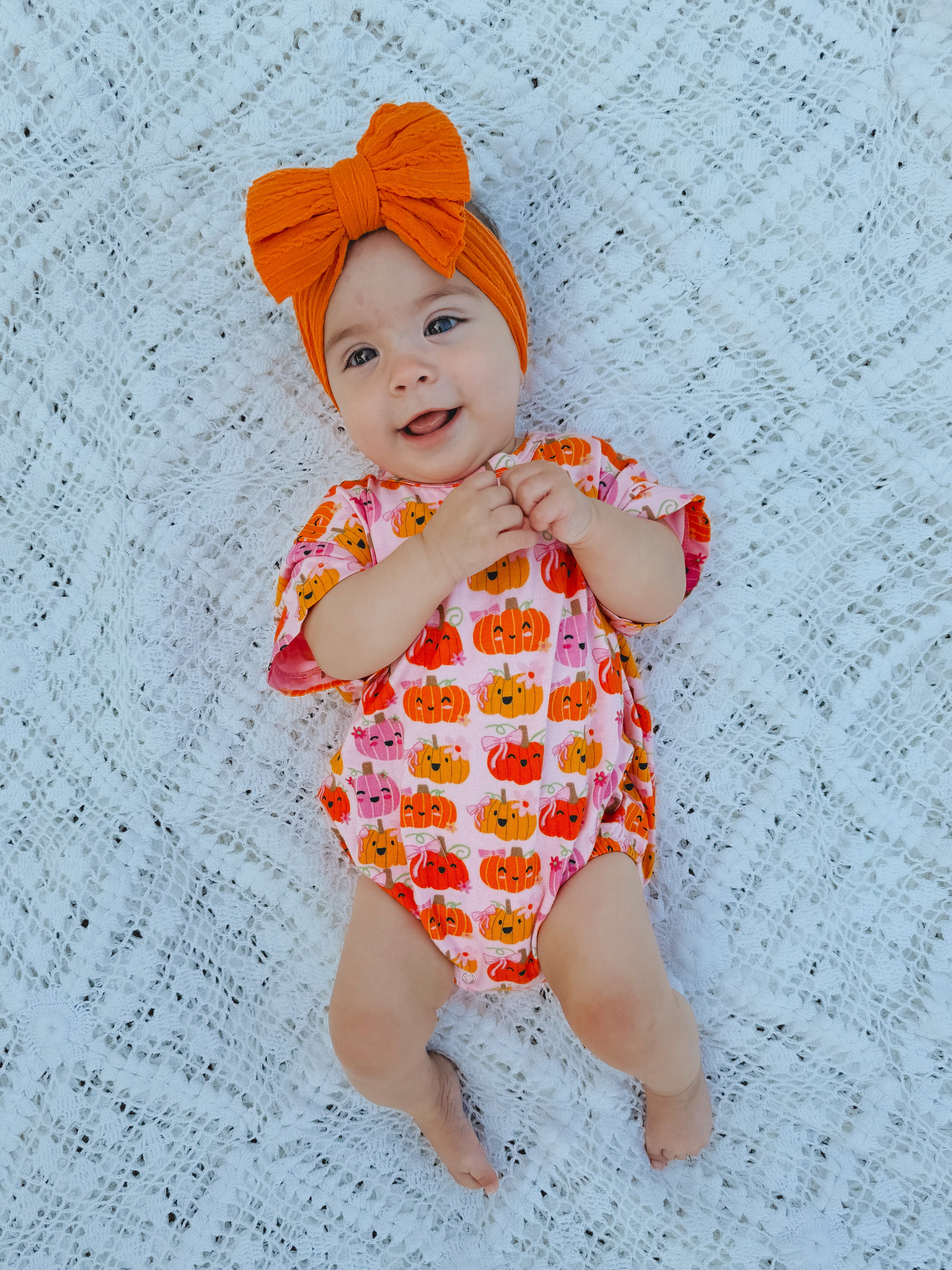 Baby lying on a white lace blanket, wearing a pumpkin-patterned outfit and an orange bow headband, smiling happily.