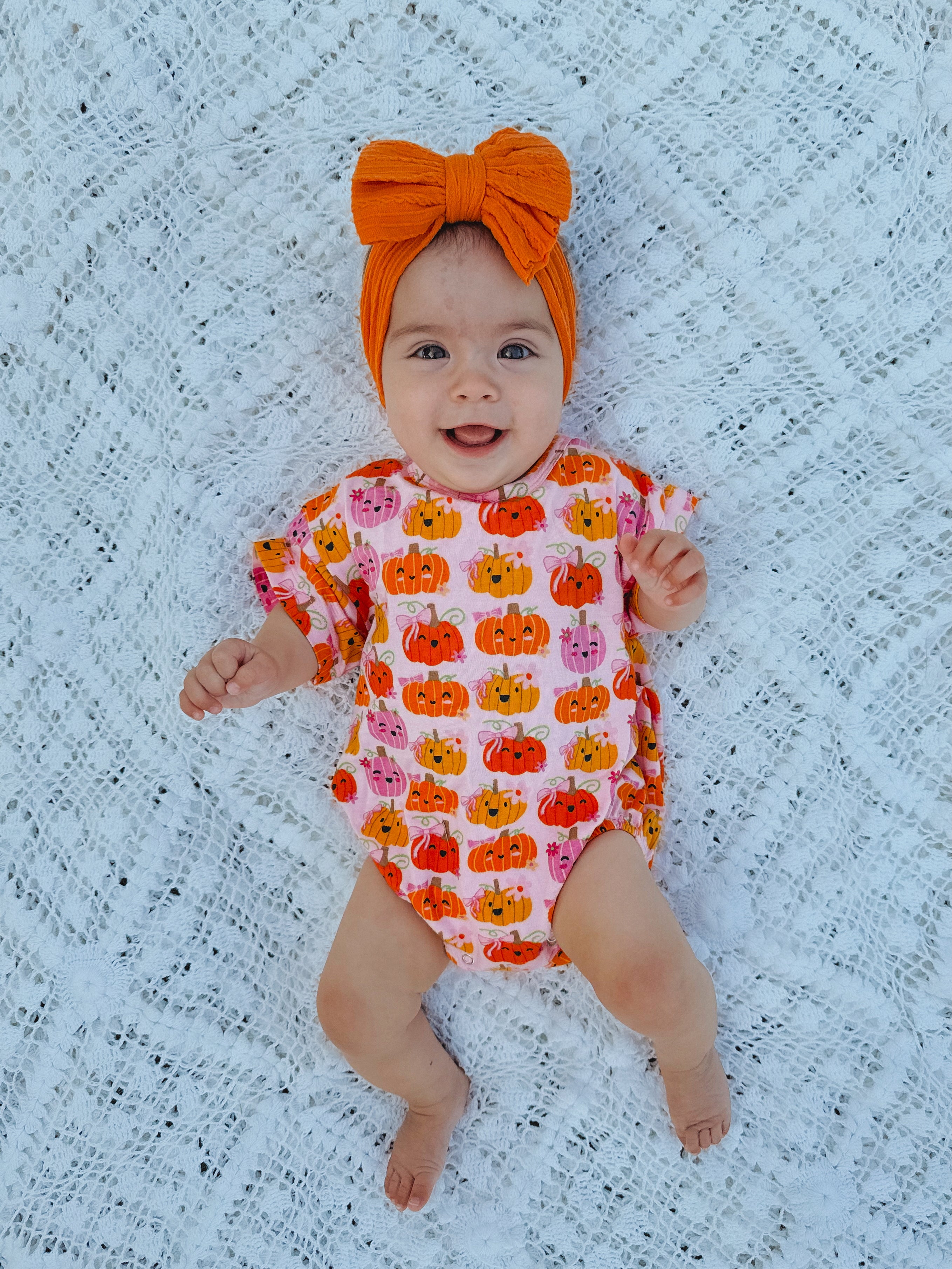Smiling baby in a pumpkin-patterned outfit with an orange bow, lying on a white lace blanket.