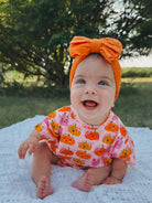 Smiling baby in a pumpkin-themed outfit and orange headband, sitting on a white blanket outdoors.