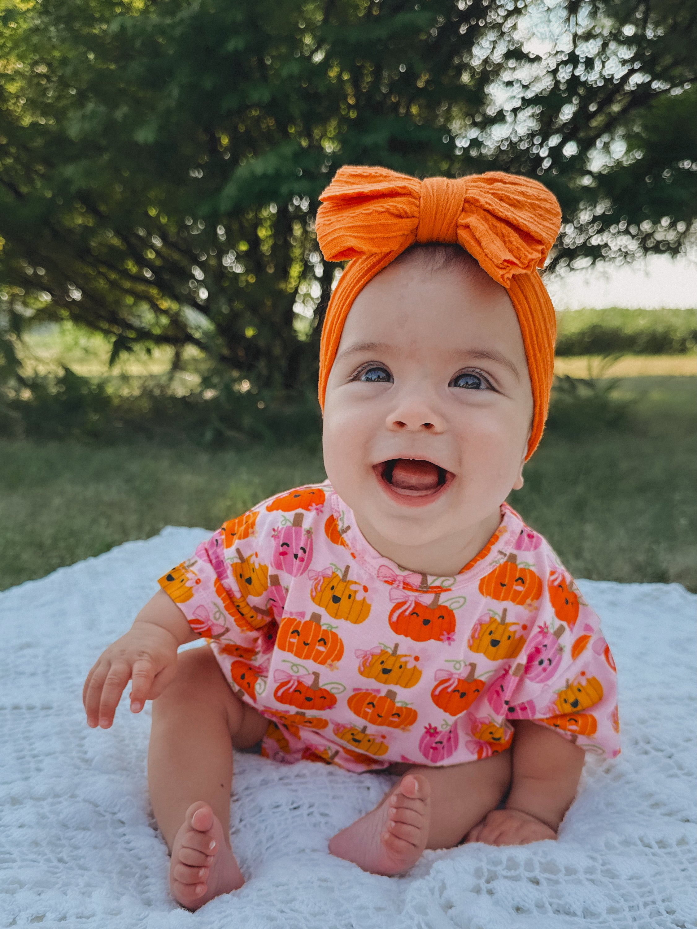 Smiling baby in a pumpkin-themed outfit and orange headband, sitting on a white blanket outdoors.