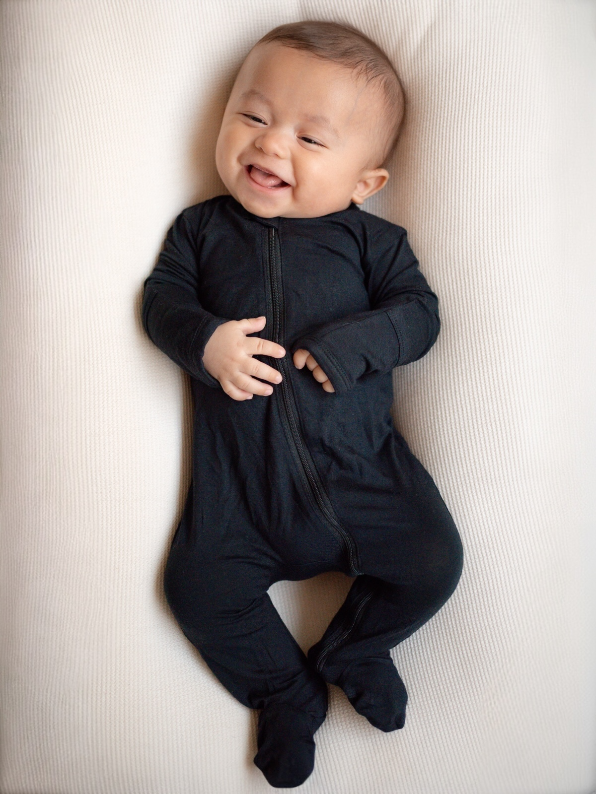 Smiling baby dressed in a black onesie, lying on a light textured surface.