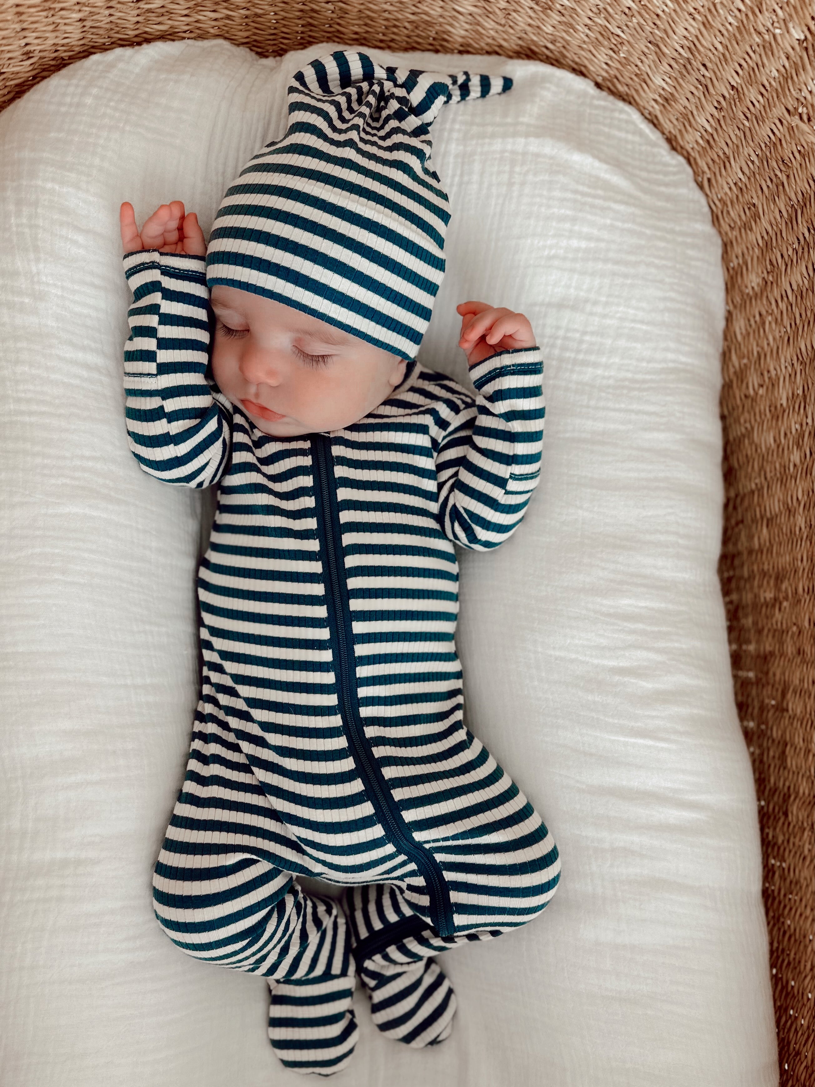 Infant sleeping on a white cushion, wearing a striped pajama and matching hat, with arms raised.