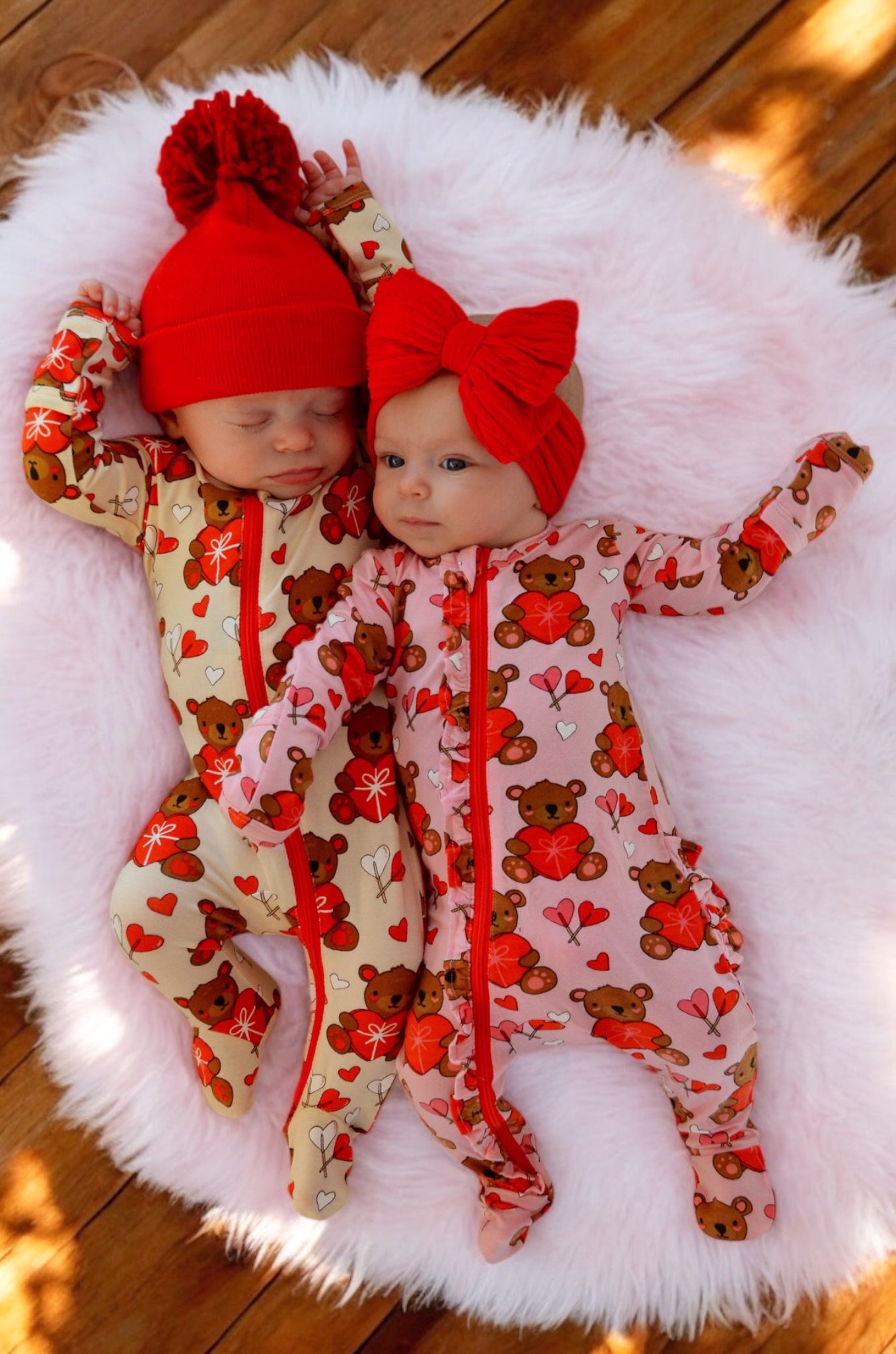 Two infants in colorful bear-patterned pajamas, one in a red hat and the other with a red bow, on a fluffy blanket.