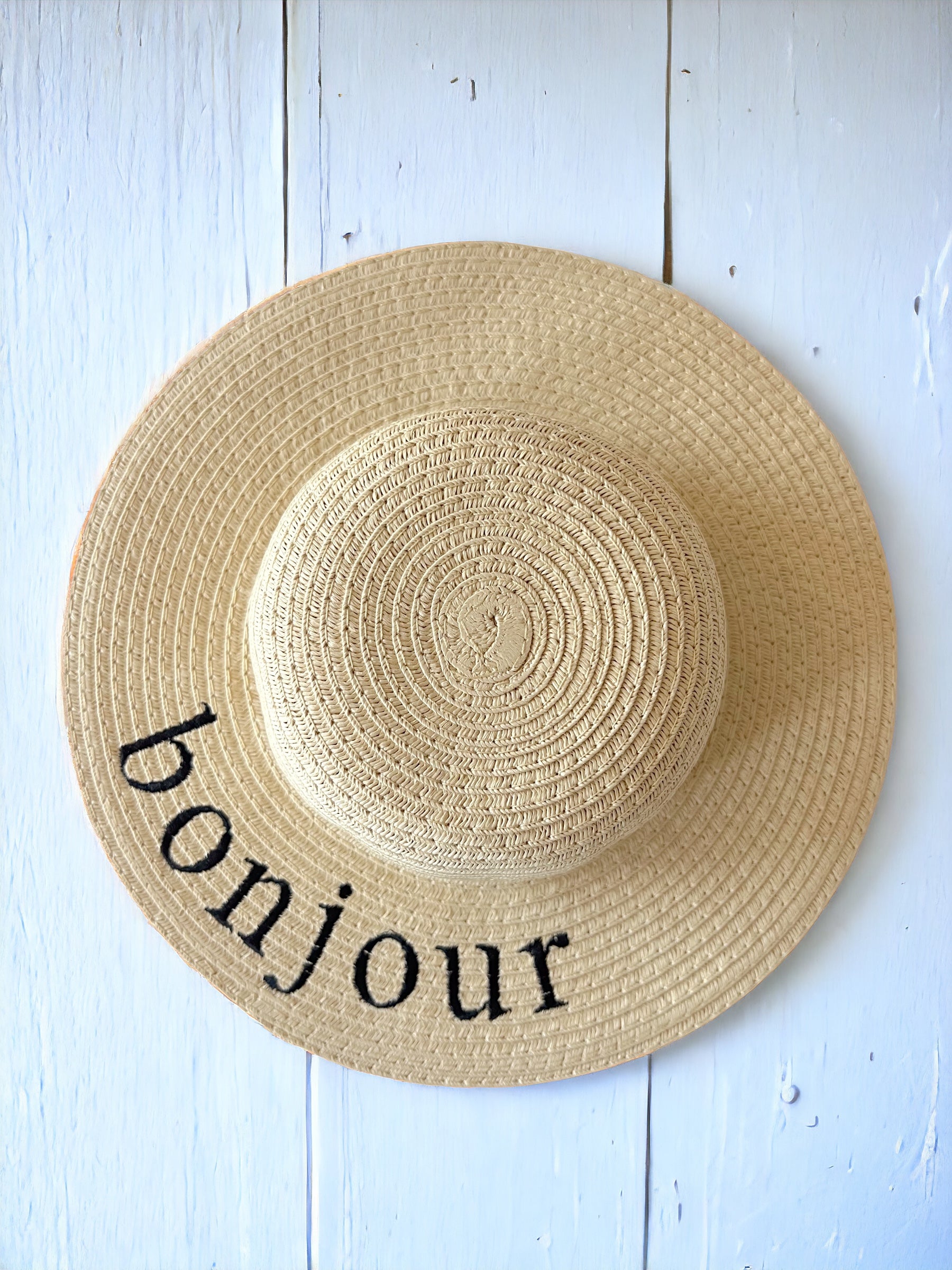 Wide-brimmed straw hat with "bonjour" embroidered in black, resting against a light wooden background.