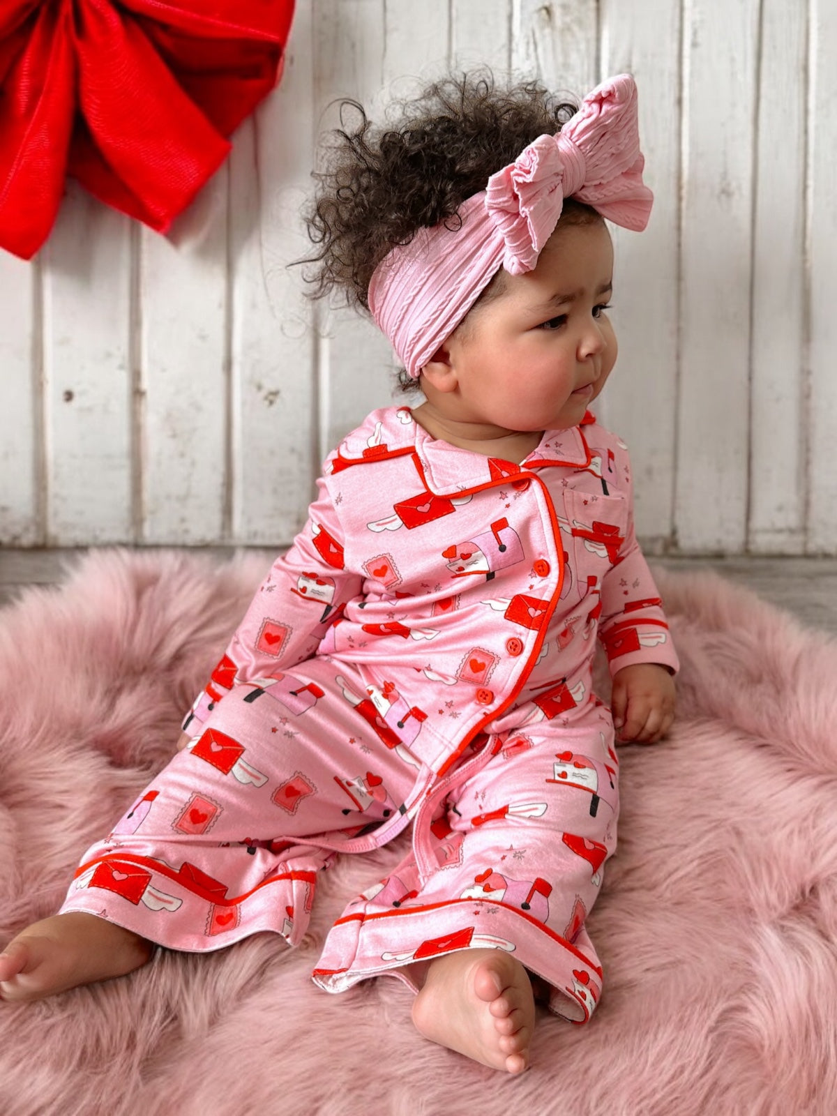 Baby in pink pajamas with festive designs, sitting on faux fur, looking to the side with a pink bow headband.
