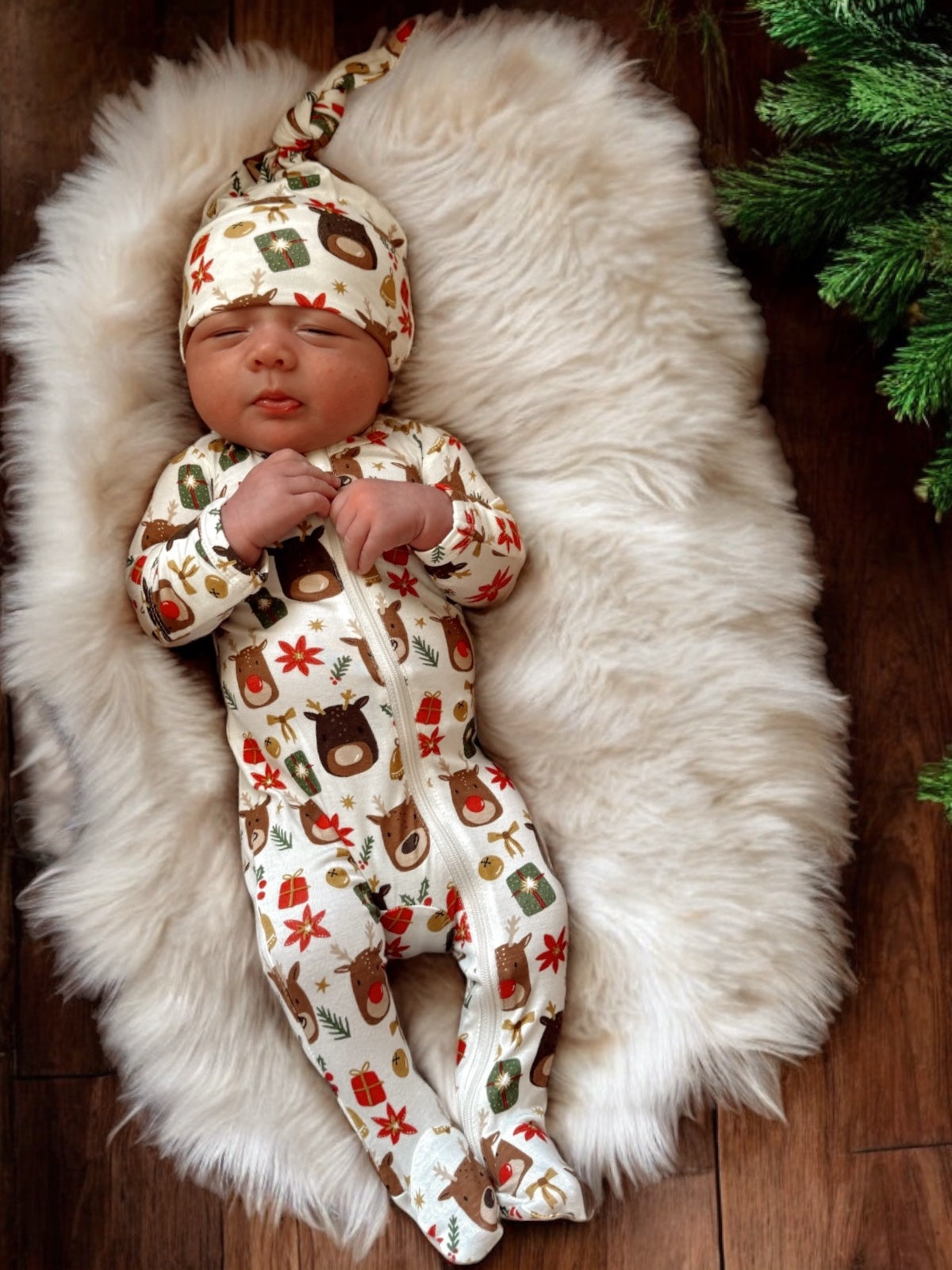 Baby wearing a festive animal-print onesie and hat, resting on a fluffy white rug with greenery nearby.
